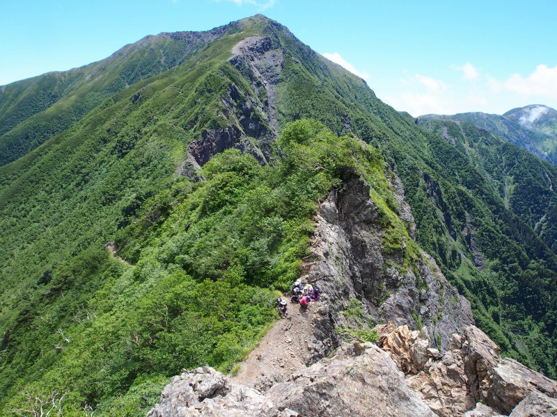 稜線上の狭い平坦地から少し登った所で振り返る。鞍部で休んでいる登山者が見えます。また、左手側の灌木帯に登山道が見えています。更にその奥に赤色チャート盤岩露出地も見えています。