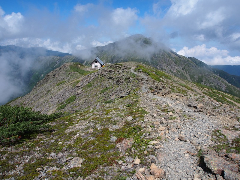 中岳山頂から荒川中岳避難小屋を写す。遠景には荒川岳（悪沢岳/東岳）が雲の合間から姿を現しています。
