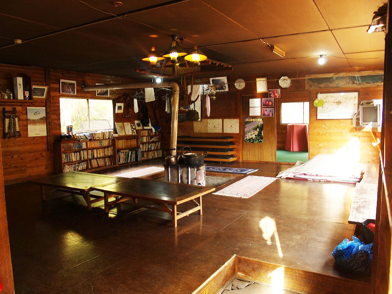オーレン小屋の談話室。中心に薪ストーブのすえられ、本棚には数多くの書籍が並べられています。 右側の低いテーブルは自炊用のテーブルとして使う事ができます。